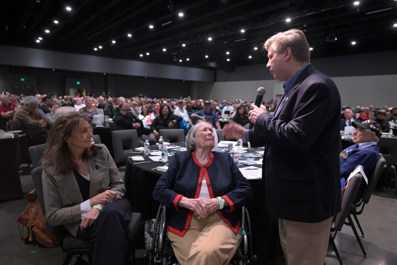 Veterans Breakfast Club15 Anniversary Celebration VBC breakfast meeting full of people with Todd holding a microphone standing next to an older woman in a chair ready to tell her veteran story