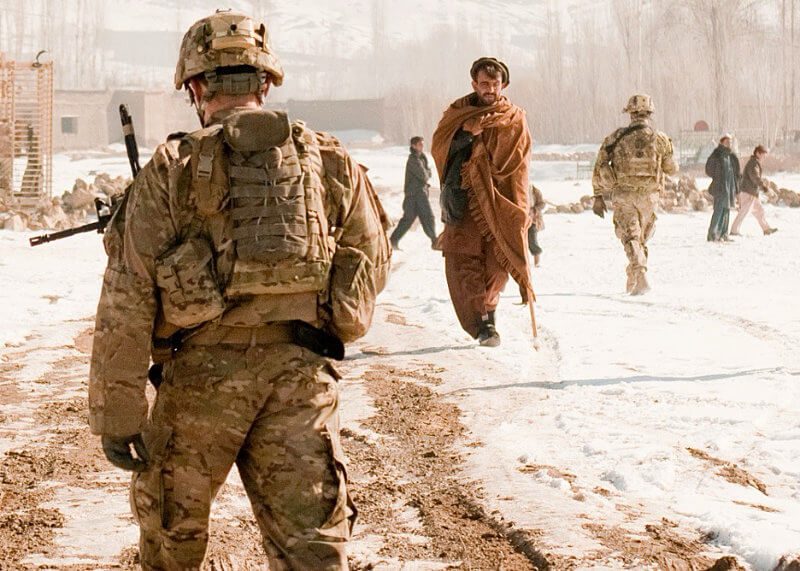 Backs of US soldiers walking down a snowy dirt road with an Afghanistan man facing the camera walking between them using a crutch